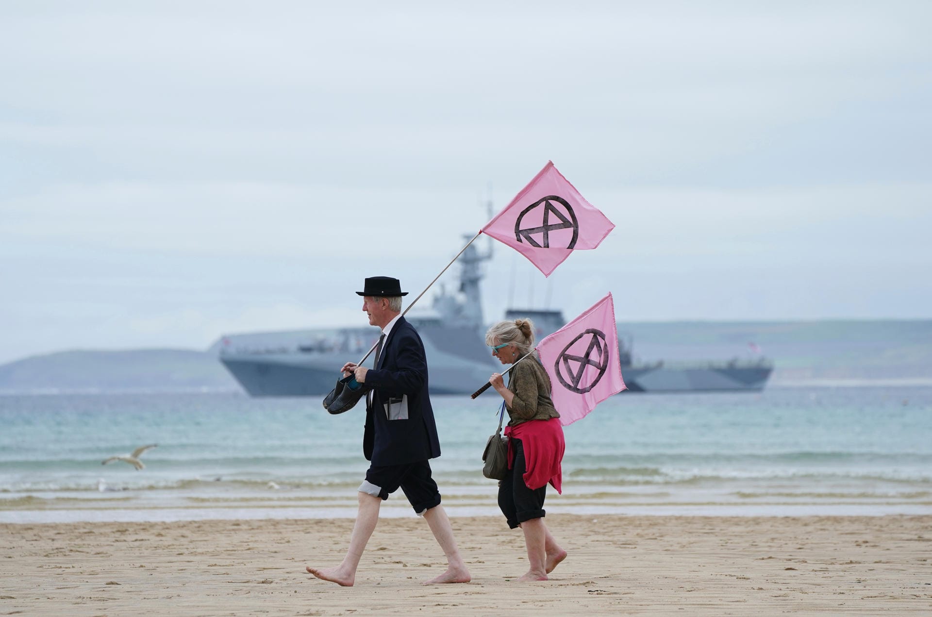 Am Strand von St Ives tragen Demonstranten zwei Fahnen mit dem Logo der Umweltbewegung Extinction Rebellion. Im Hintergrund liegt die HMS Tamar vor Anker, ein Kriegsschiff der britischen Marine. Auch der neueste Flugzeugträger der Royal Navy, die HMS Prince of Wales (nihct im Bild) gehört zum Sicherheitskonzept des G7-Gipfels in Cornwall.