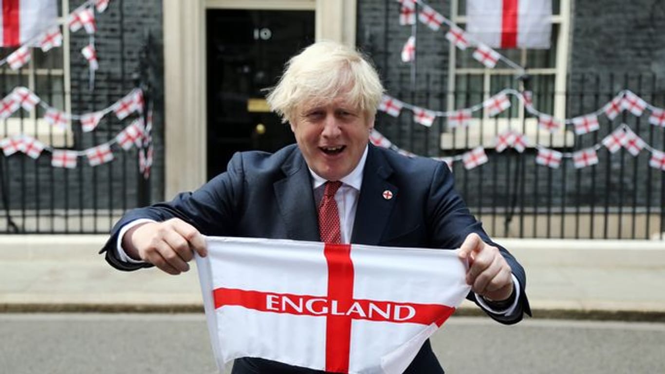 Premierminister Boris Johnson hält eine englische Fahne vor sich in die Höhe.