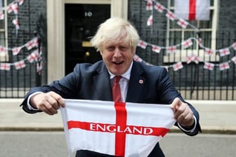 Premierminister Boris Johnson hält eine englische Fahne vor sich in die Höhe.