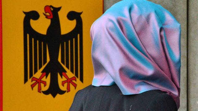 Eine junge Frau mit Kopftuch geht an einem Behördenschild mit dem Bundesadler vorbei.