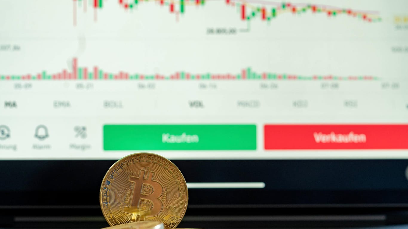 Eine Bitcoin-Münze vor einem Bildschirm mit fallenden Kursen (Symbolbild): Der Wert der Kryptowährung sackt unter 30.000 US-Dollar ab.