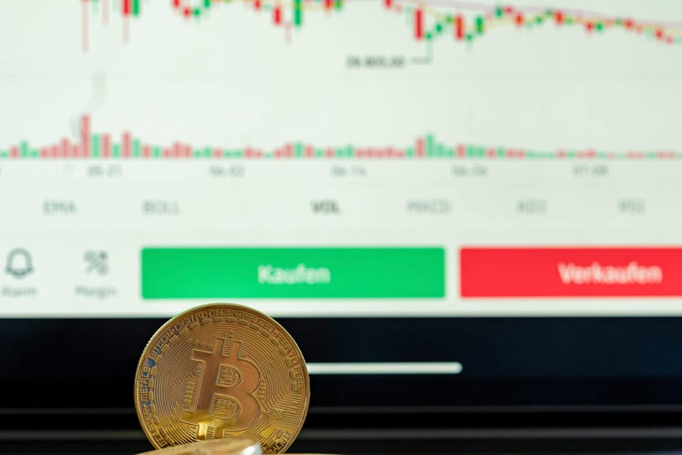 Eine Bitcoin-Münze vor einem Bildschirm mit fallenden Kursen (Symbolbild): Der Wert der Kryptowährung sackt unter 30.000 US-Dollar ab.