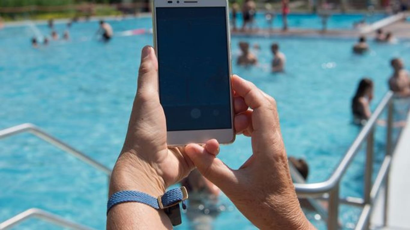 Eine Frau hält im Freibad ein Smartphone in Richtung des Pools