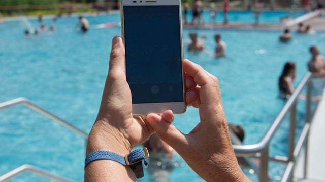 Eine Frau hält im Freibad ein Smartphone in Richtung des Pools