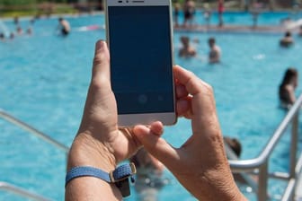 Eine Frau hält im Freibad ein Smartphone in Richtung des Pools