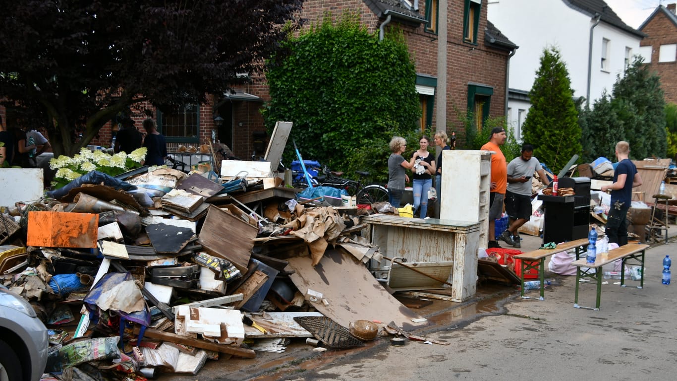 Elke K. steht mit Nachbarn und Helfern vor ihrem Haus. Im Vordergrund türmen sich unbrauchbare Möbel und Müll.