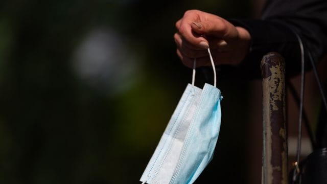 Ein Frau hält vor einem Bäckereigeschäft ihren Mundschutz in der Hand.