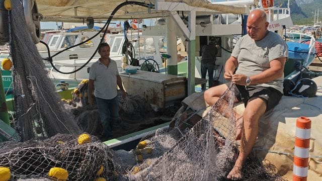 Der Leiter der Fischerei-Kooperative in Antalya, Cengiz Balta, steht auf einem Boot, während ein weiteres Kooperativen-Mitglied im Hafen ein Netz flickt.
