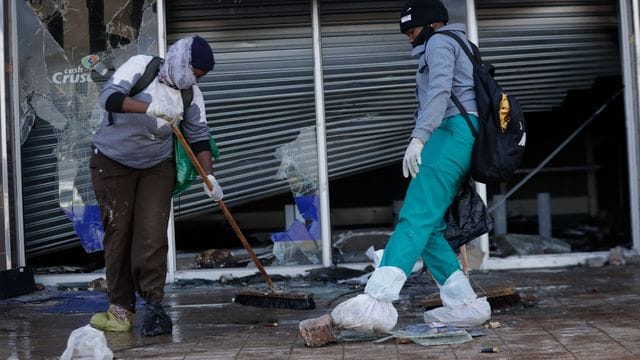 Zwei Personen räumen in Vosloorus (Südafrika) nach Ausschreitungen vor einem zerstörten Einkaufszentrum den Gehweg auf.
