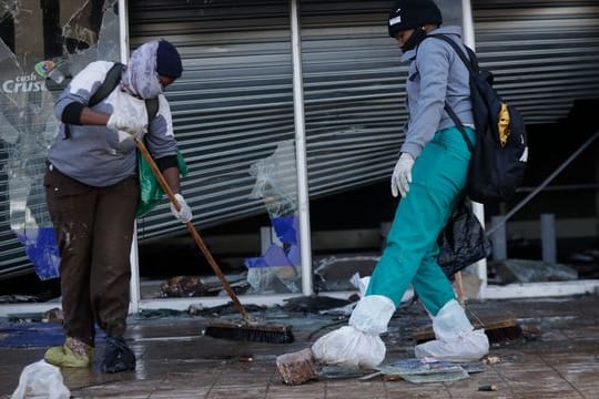Zwei Personen räumen in Vosloorus (Südafrika) nach Ausschreitungen vor einem zerstörten Einkaufszentrum den Gehweg auf.