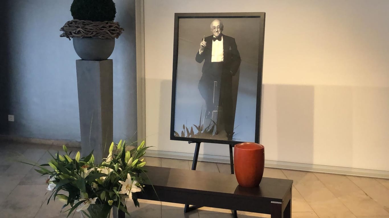 Ein Foto von Alfred Biolek, auf dem er freundlich lächelnd den Zeigefinger hebt, davor die Urne des Entertainers und seine Lieblingsblumen, weiße Lilien: In einem Kölner Bestattungsunternehmen können Trauernde Abschied von dem Verstorbenen nehmen.