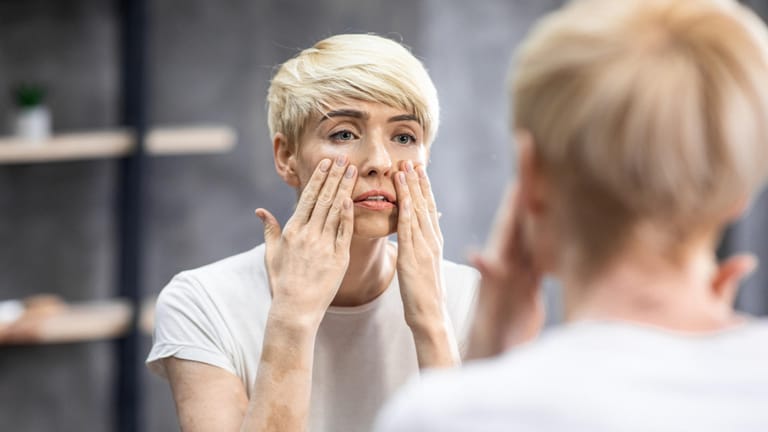 Eine Frau betrachtet ihr Gesicht kritisch im Spiegel. Die Hautalterung ist ein schleichender Prozess, der schon mit Mitte Zwanzig beginnt.