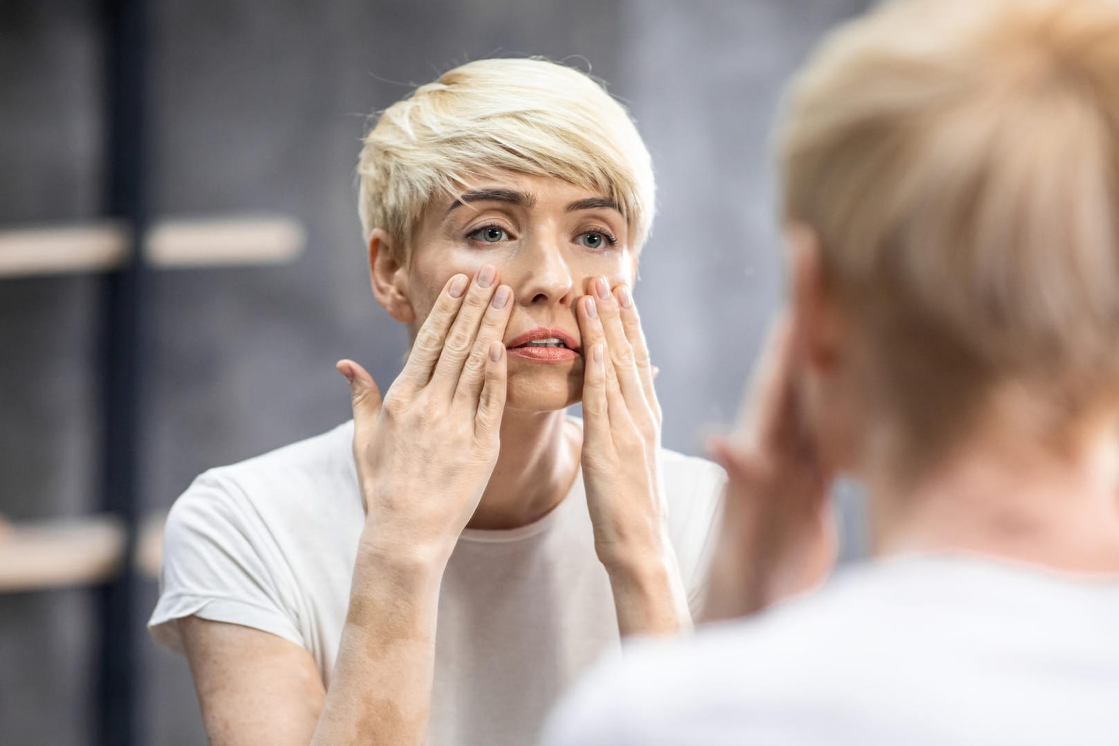 Eine Frau betrachtet ihr Gesicht kritisch im Spiegel. Die Hautalterung ist ein schleichender Prozess, der schon mit Mitte Zwanzig beginnt.