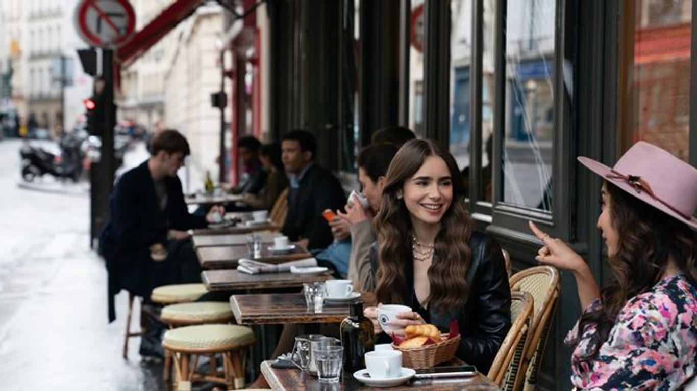 Emily (Lily Collins, m) sitzt mit einer Freundin im Café - Filmszene aus der ersten Staffel der Netflix-Serie "Emily in Paris".