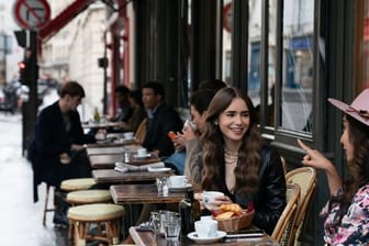 Emily (Lily Collins, m) sitzt mit einer Freundin im Café - Filmszene aus der ersten Staffel der Netflix-Serie "Emily in Paris".