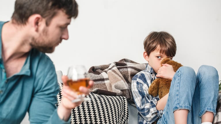 Ein Vater trinkt Alkohol, sein Sohn sitzt verschüchtert neben ihm: Für Kinder suchtkranker Menschen gibt es spezielle Angebote.