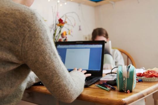 Eine Mutter arbeitet am Küchentisch während ihre Tochter gegenüber in ein Tablet guckt.