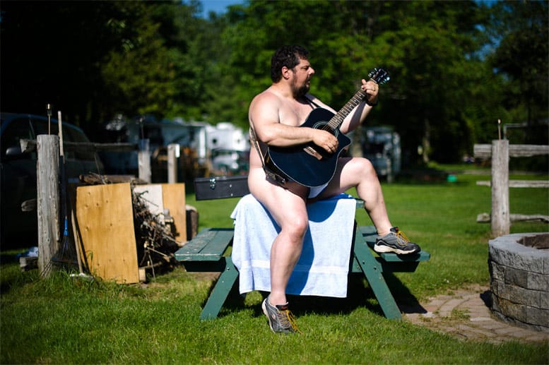 Bedeckt mit seiner Gitarre und Turnschuhen an den Füßen stimmt ein musikalischer Anhänger der Freikörperkultur auf einer Wiese im Bare Oaks Family Naturist Park ein Liedchen an.