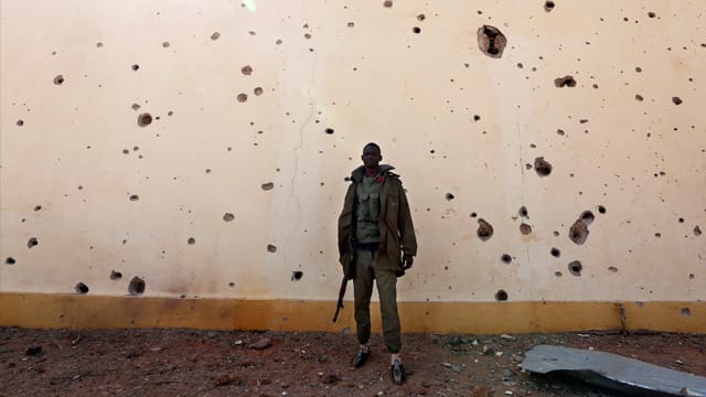 Ein Soldat steht in Konna vor einer von Kugeln und Bombensplittern durchlöcherten Wand. Ein Soldat steht in Konna vor einer von Kugeln und Bombensplittern durchlöcherten Wand.