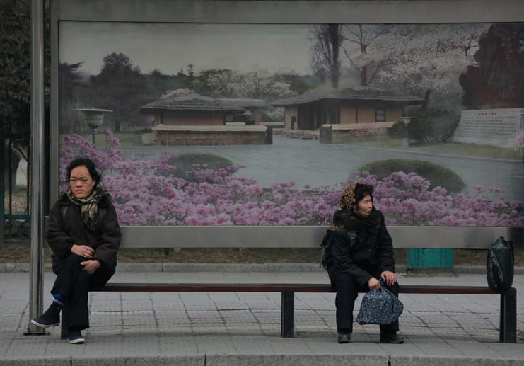 Zwei Frauen warten an einer Bushaltestelle in der Hauptstadt auf den Bus. Das Plakat im Hintergrund zeigt eine idyllische Landschaft