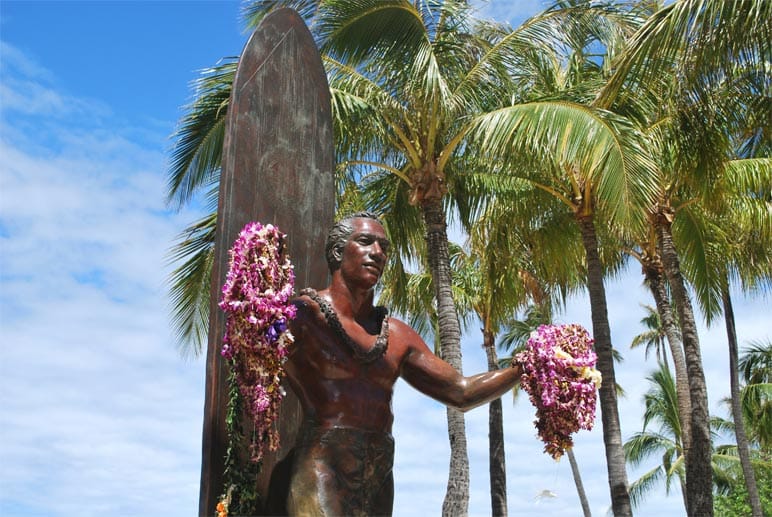 Eine mit einem frischen Blütenkranz (Lei) verzierte Bronzestatue erinnert an Duke Kahanamoku, den Erfinder des modernen Wellenreitens und Godfather aller Surfer.