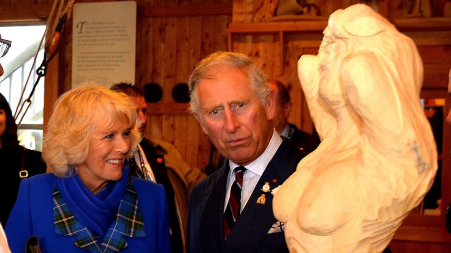 Das royale Paar bewundert auf seiner Reise eine hölzerne Gallionsfigur. Prinz Charles schaut etwas entsetzt auf die blanken Brüste der Figur, Camilla freut sich.
