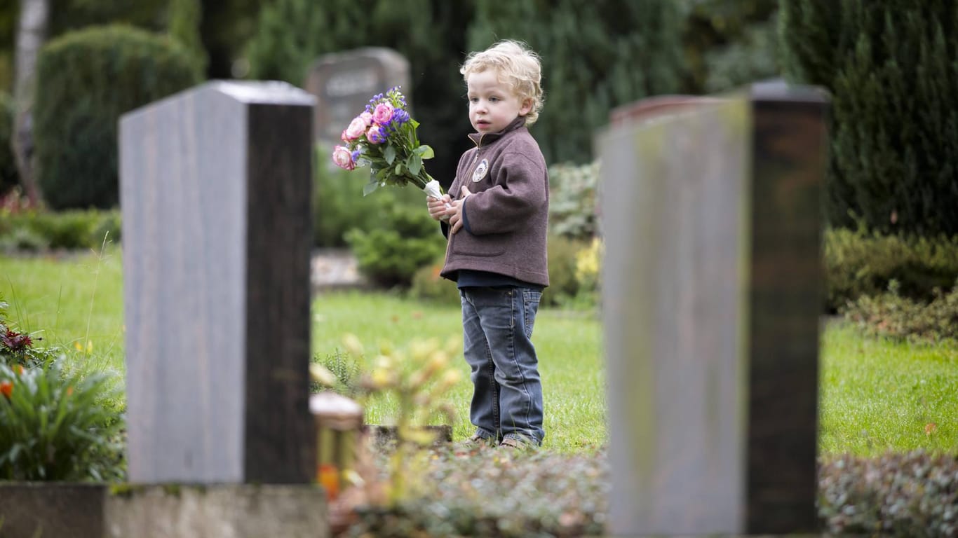 Ein dreijähriger Junge steht mit Blumen an einem Grab. Kinder haben eine eigene Art zu trauern.