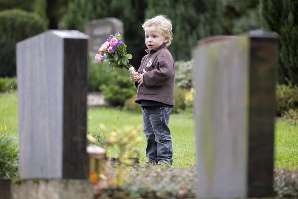 Ein dreijähriger Junge steht mit Blumen an einem Grab. Kinder haben eine eigene Art zu trauern.