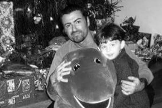James Kennedy als Kind zusammen mit George Michael auf einem frühen Foto, das die beiden vor einem Weihnachtsbaum und Geschenken zeigt.