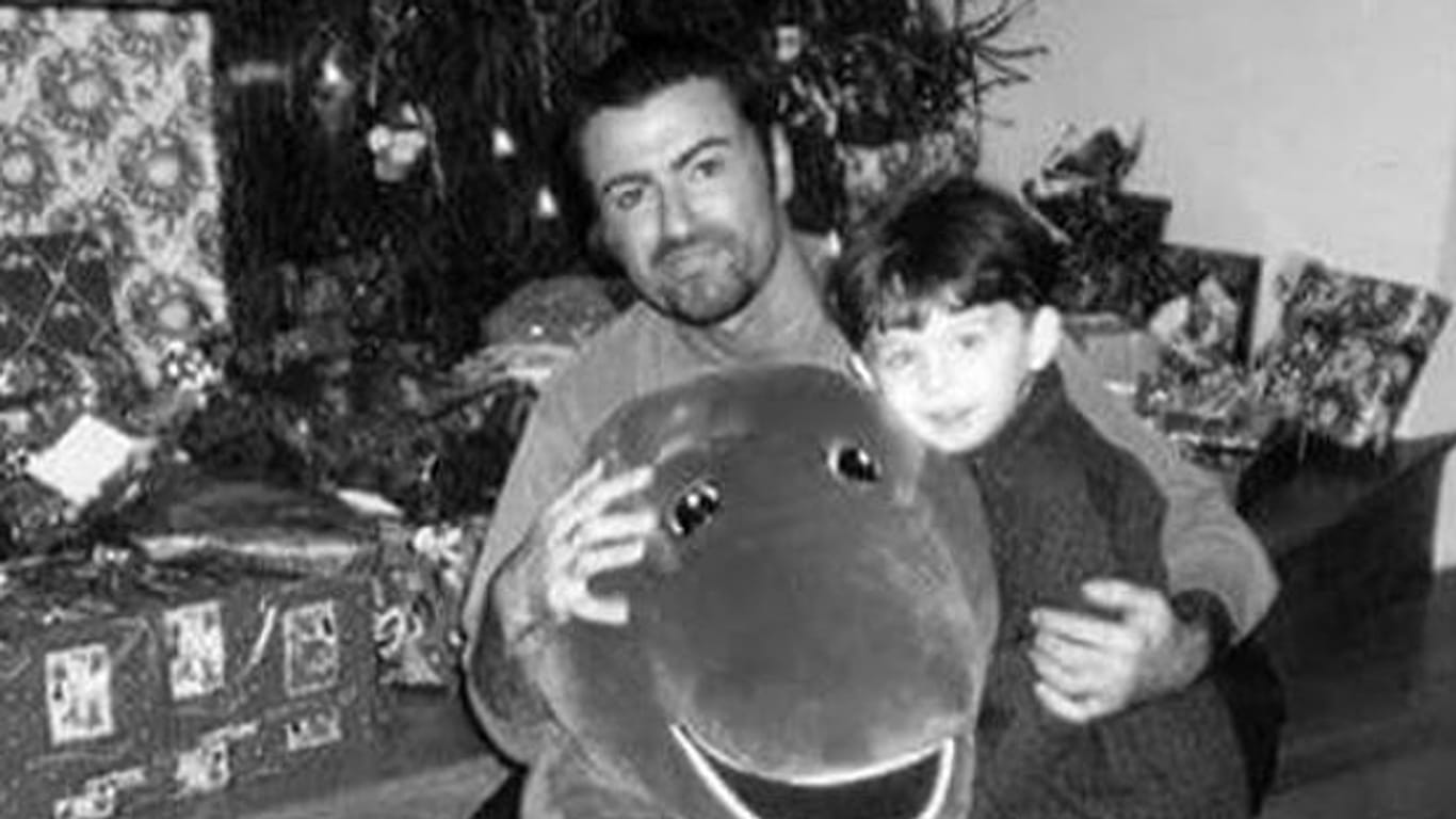 James Kennedy als Kind zusammen mit George Michael auf einem frühen Foto, das die beiden vor einem Weihnachtsbaum und Geschenken zeigt.