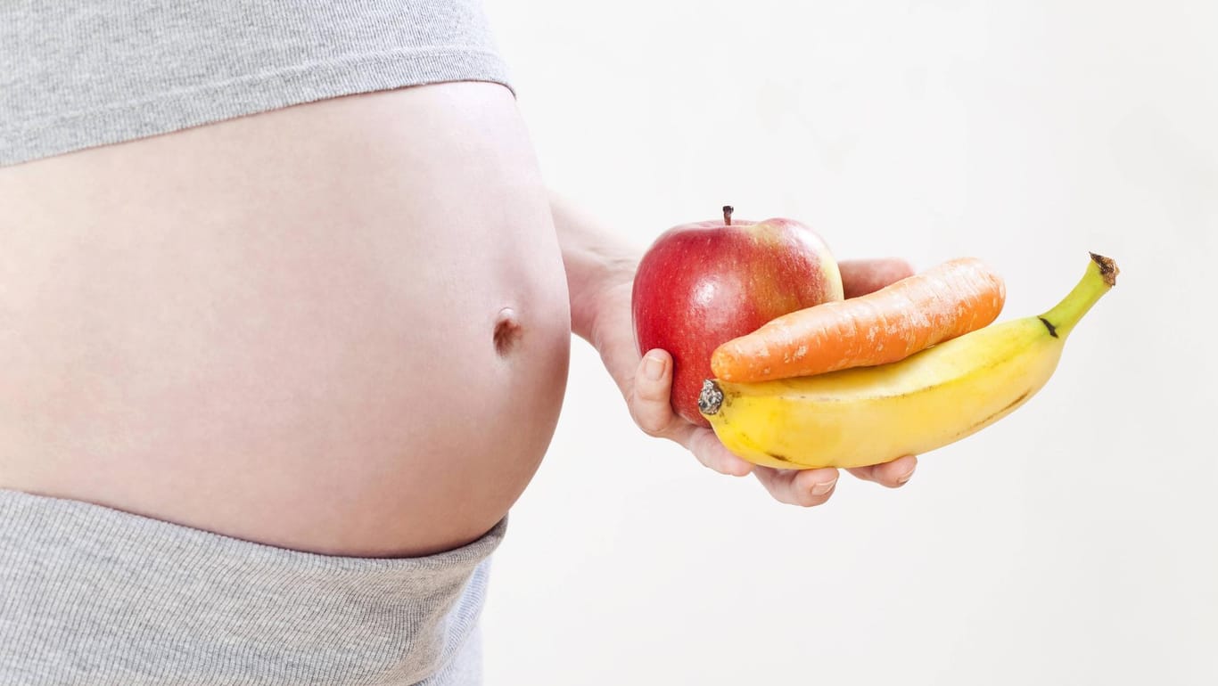 Schwangere Frau hält Apfel, Banane und eine Karotte in der Hand.