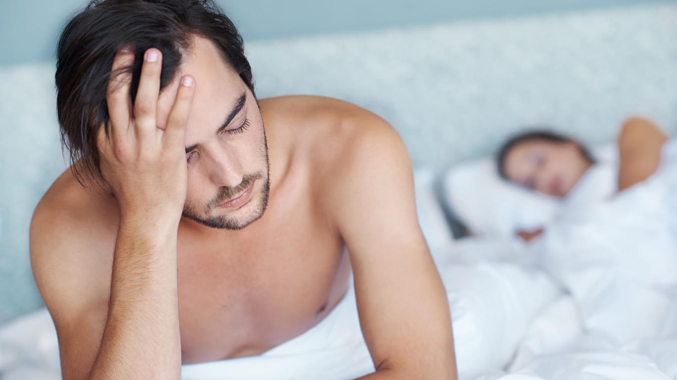 Junger Mann sitzt auf dem Bett während seine Frau im Hintergrund schläft.