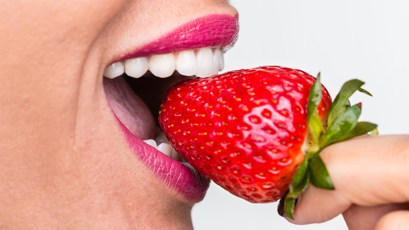 Eine Frau beißt in eine Erdbeere: Die süßen roten Früchte sind gesund – doch nicht als Paste auf den Zähnen. Der Fruchtzucker und die Säure von Früchten schaden dem Zahnschmelz.