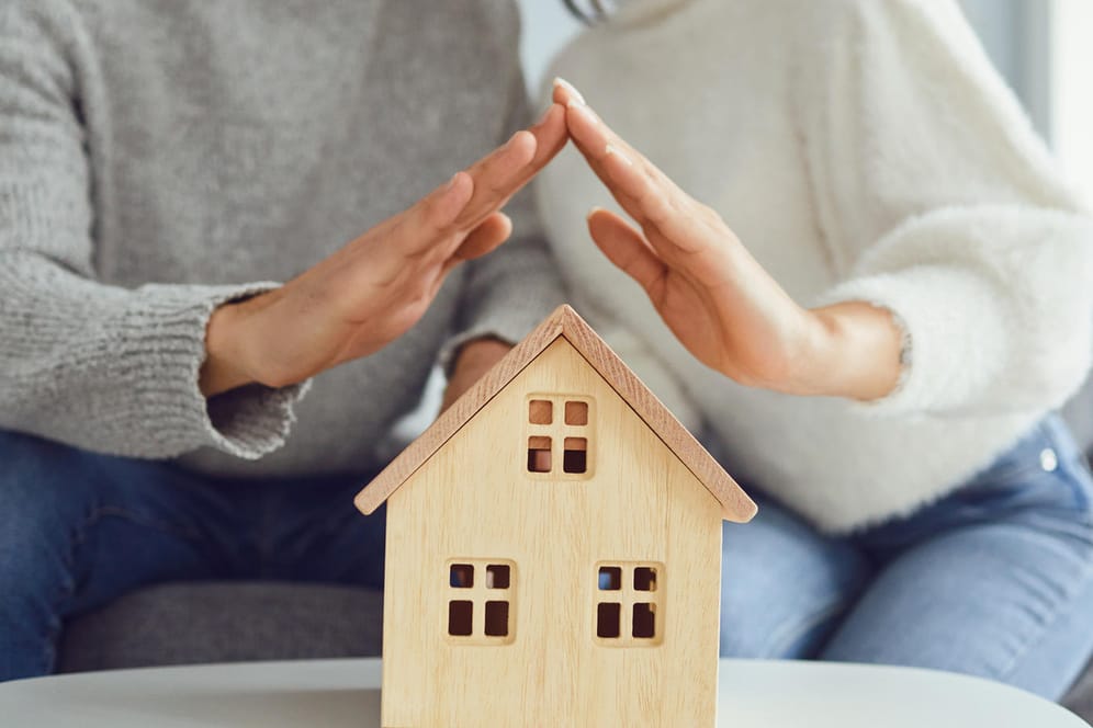 Ein Paar hält seine Hände wie ein Dach über ein Modell eines Hauses (Symbolbild): Wenn der Bausparvertrag zuteilungsreif ist, kann es endlich losgehen mit dem Hauskauf.