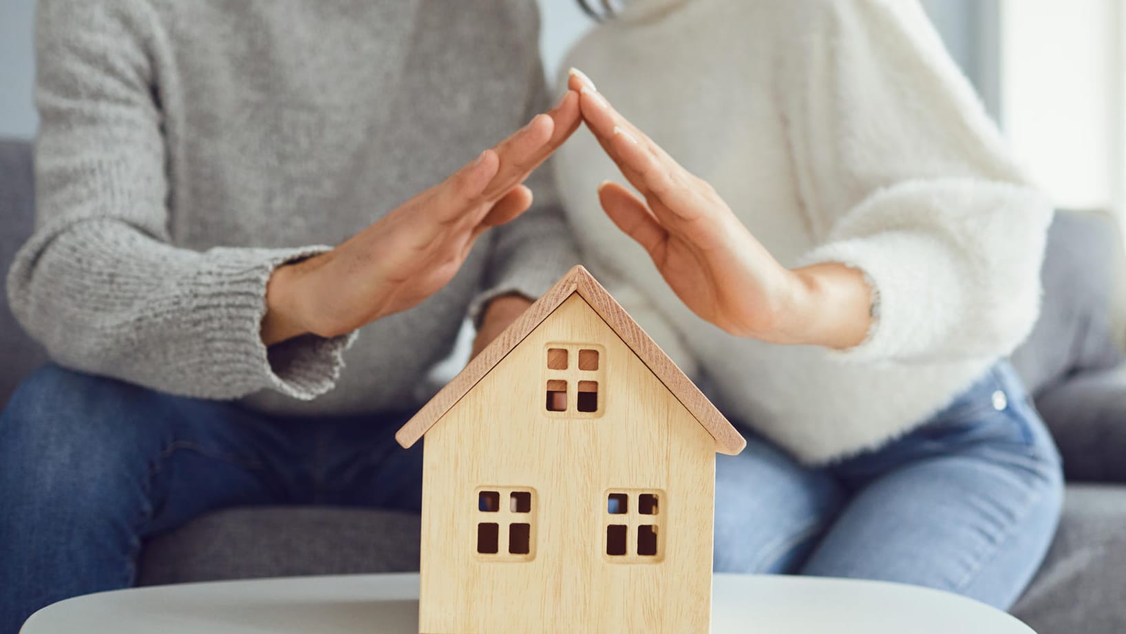 Ein Paar hält seine Hände wie ein Dach über ein Modell eines Hauses (Symbolbild): Wenn der Bausparvertrag zuteilungsreif ist, kann es endlich losgehen mit dem Hauskauf.