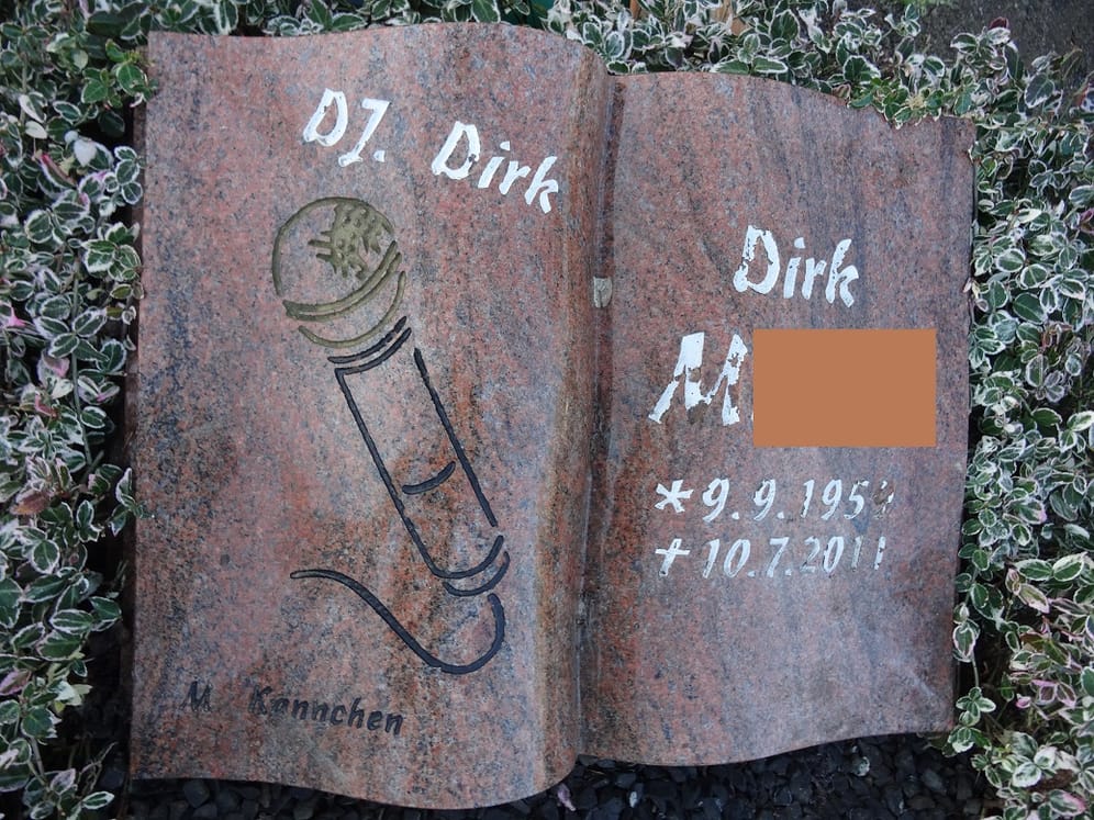 DJ-Dirk-Grabstein: Das in Stein gemeißelte Mikrofon und die Abkürzung für Discjockey (DJ) auf dem Grabmal verweisen darauf, was den Toten besonders begeisterte.