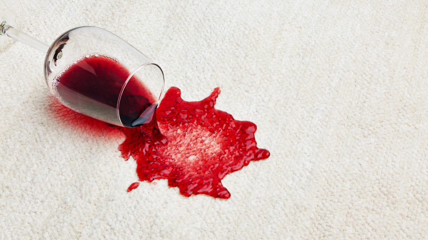 Ein Rotweinglas ist auf dem hellen Teppich ausgekippt.