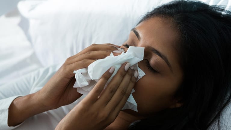 Erkältete Frau liegt im Bett und putzt sich die Nase mit einem Taschentuch.
