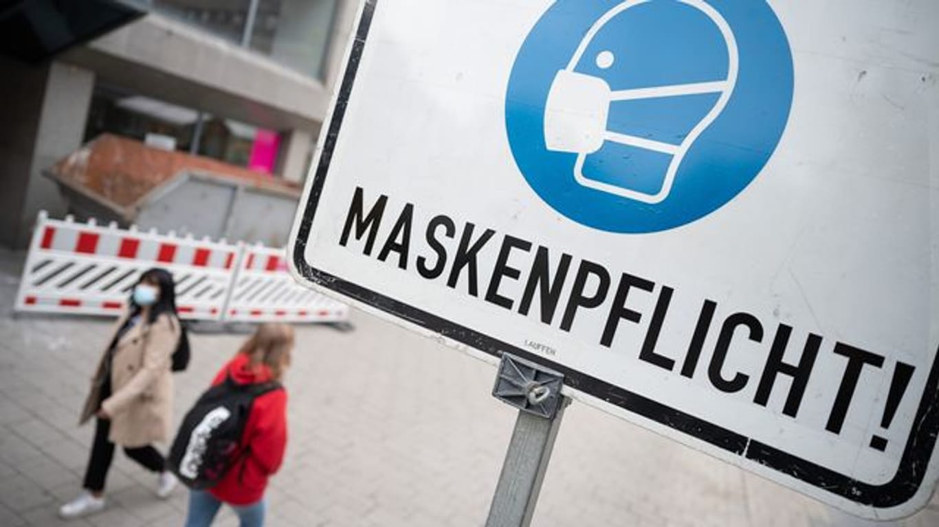 Zwei Passantinnen gehen in einer Fußgängerzone hinter einem Schild vorbei, auf dem "Maskenpflicht" steht.