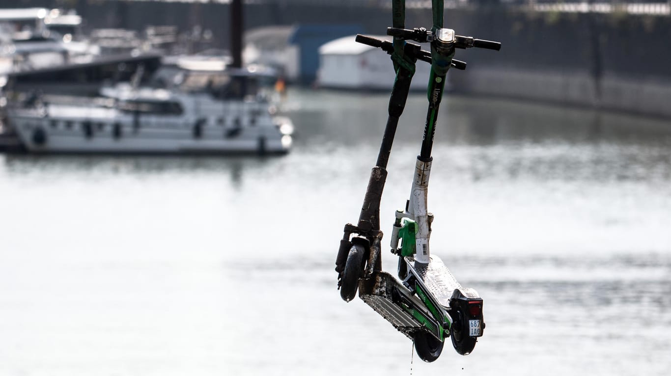 Zwei E-Scooter hängen an einem Kran: Spezialtaucher, ein Beiboot und ein Autokran sollen die zahlreichen E-Scooter aus dem Rheinauhafen bergen – die Ausbeute ist bisher mau.