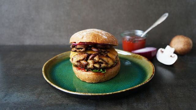 Pilzburger: Braune Champignons und Dinkel-Bulgur liegen zusammen mit Cheddar und roten Zwiebelringen zwischen zwei Brötchenhälften.