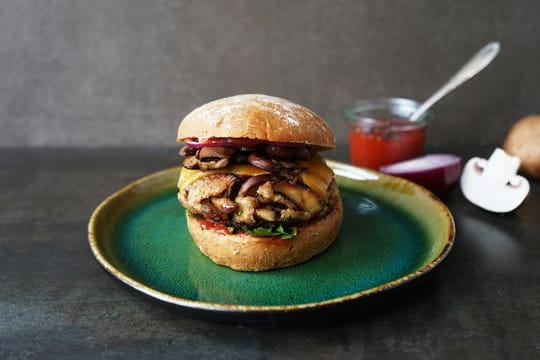 Pilzburger: Braune Champignons und Dinkel-Bulgur liegen zusammen mit Cheddar und roten Zwiebelringen zwischen zwei Brötchenhälften.