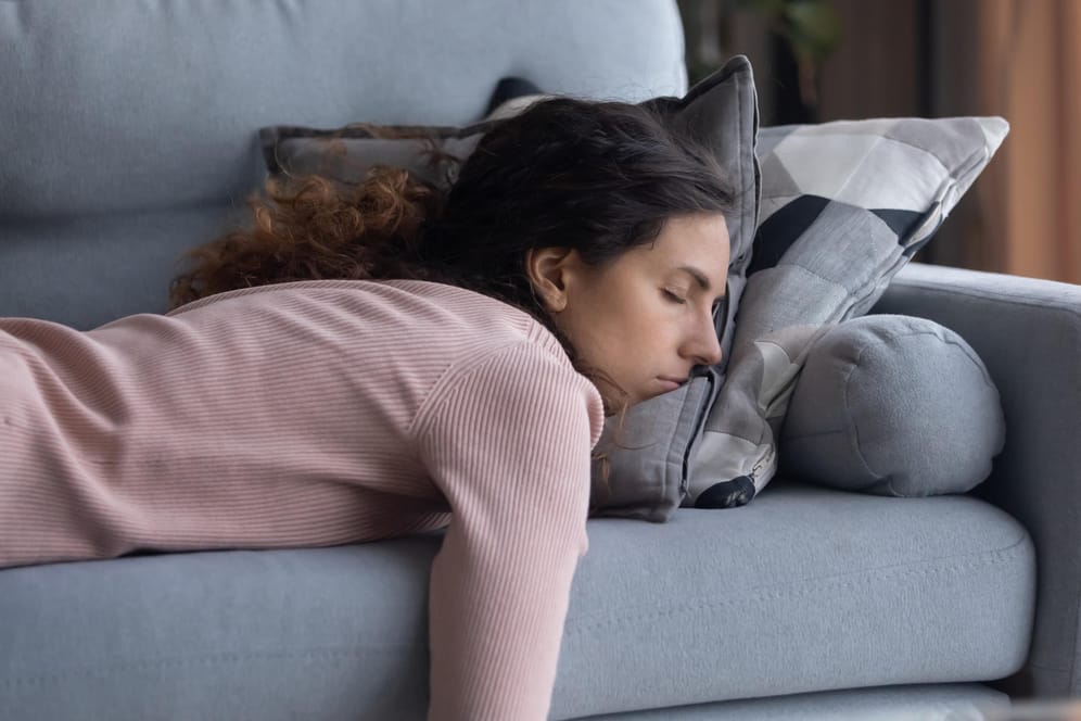 Eine junge Frau liegt erschöpft auf dem Sofa. Chronische Müdigkeit kann von Dauerstress kommen und ist ein Warnsignal des Körpers.