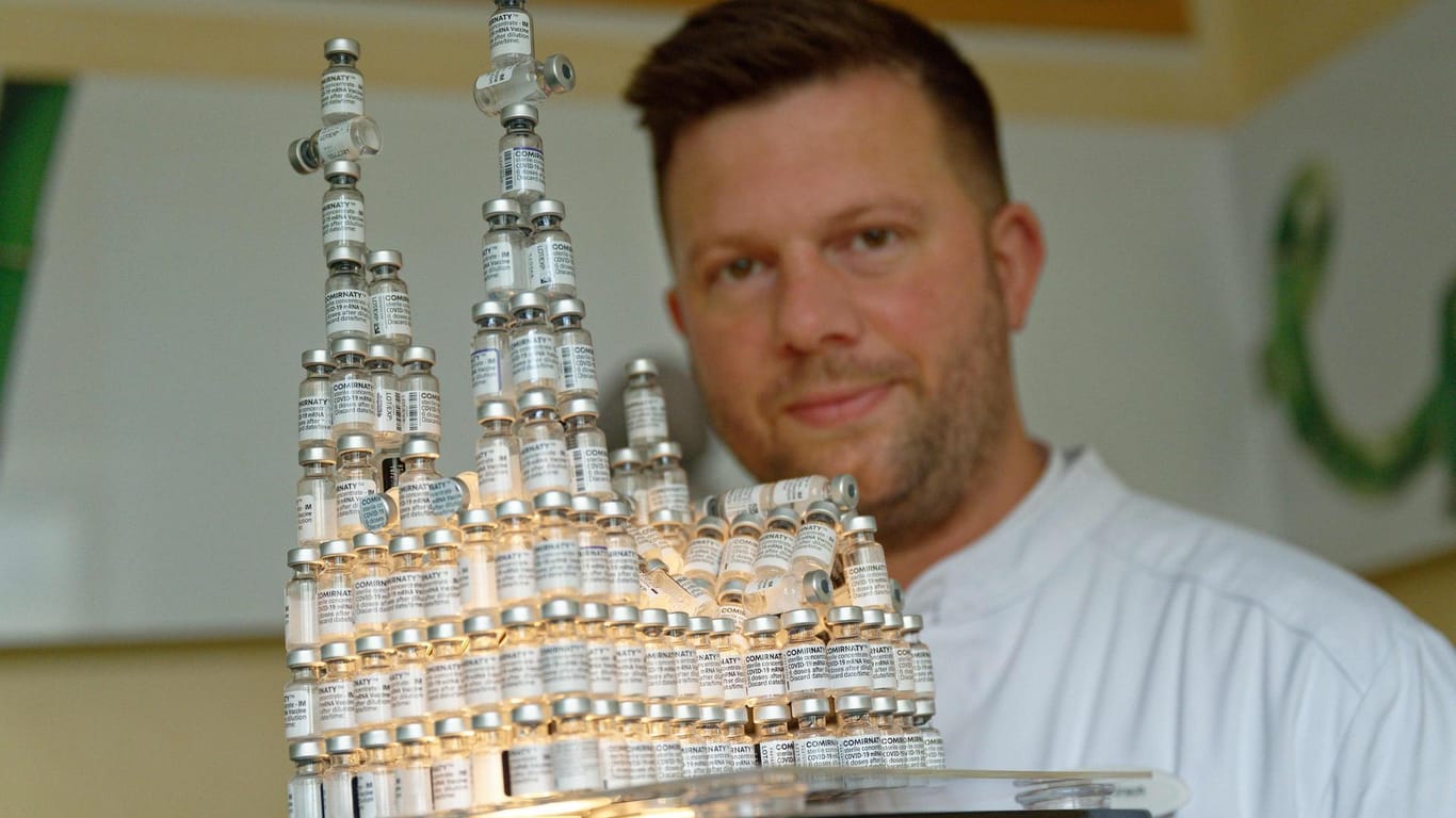 Der Kölner Arzt Bastian Kirsch sitzt neben einem Modell des Kölner Doms: Dieses hat er aus 220 leeren Impfflaschen gebaut.