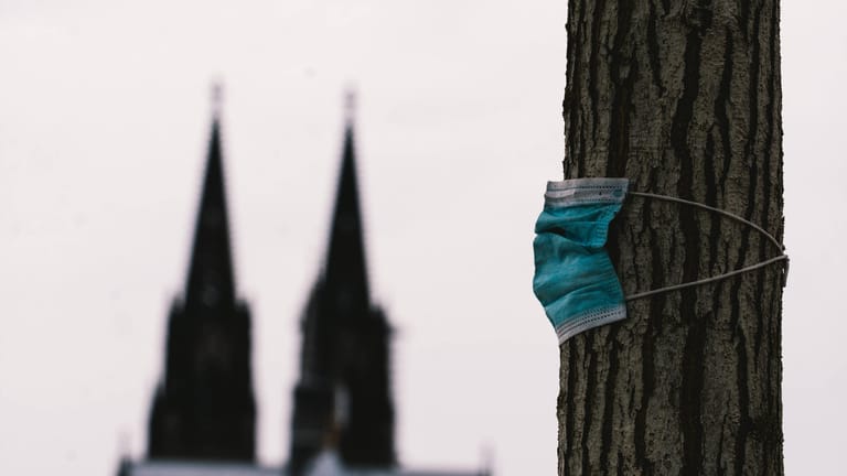 Eine OP-Maske hängt an einem Baum, im Hintergrund der Kölner Dom: In der Stadt am Rhein sieht man die Zeit für den Wegfall aller Maßnahmen noch nicht gekommen.