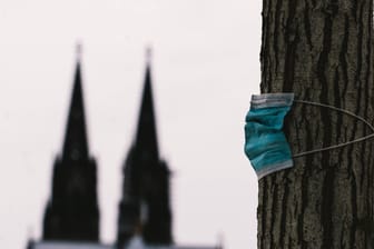 Eine OP-Maske hängt an einem Baum, im Hintergrund der Kölner Dom: In der Stadt am Rhein sieht man die Zeit für den Wegfall aller Maßnahmen noch nicht gekommen.