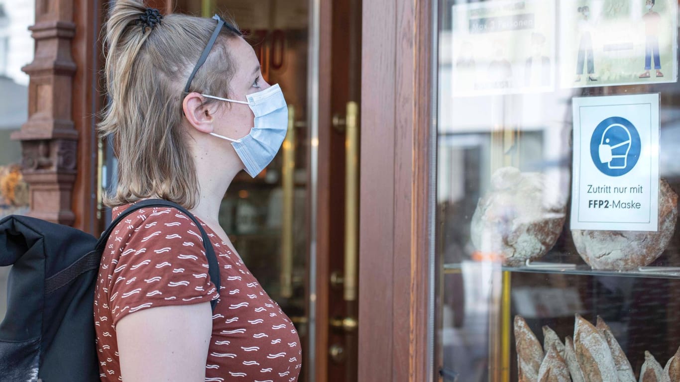 Eine Frau mit Maske steht an einer Bäckerei (Symbolbild). Der Inzidenzwert in Deutschland ist derzeit volatil.