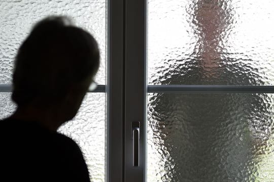 Eine Frau steht an einer Glasscheibe, hinter der die Silhouette eines Mannes zu sehen ist (Symbolbild): In Bayern gelten bald härtere Strafen für Stalker.