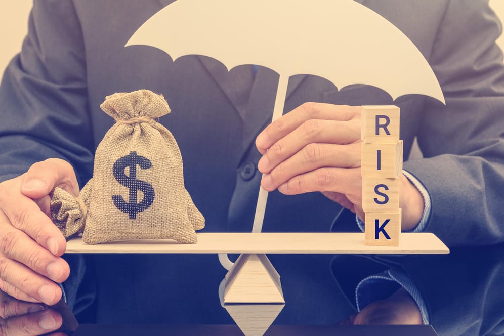 Geschäftsmann hält einen weißen Schirm schützend über eine ausbalancierte Waage auf der ein Sack Geld und Holzwürfel mit der Aufschrift Risiko stehen