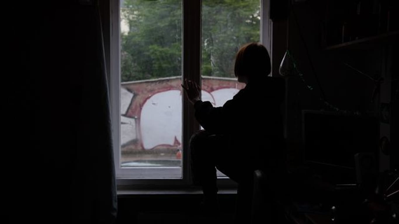Eine Jugendliche sitzt in einem abgedunkelten Zimmer am Fenster.